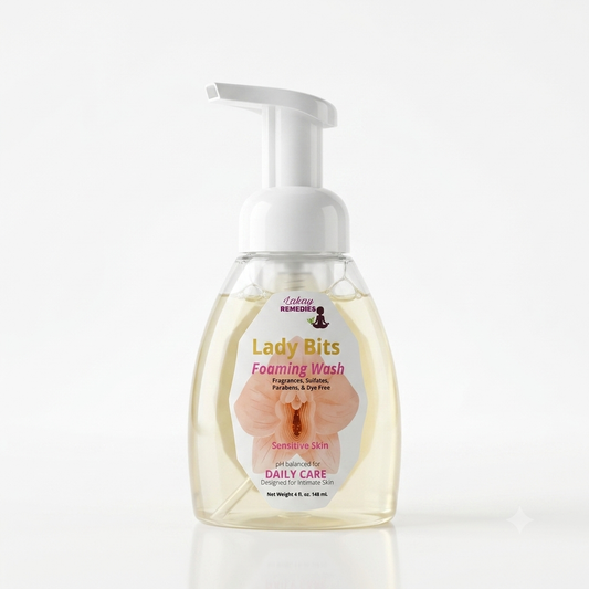 A bottle of Lady Bits Foaming Wash, featuring a clear plastic bottle filled with pale yellow liquid, a white pump dispenser, and a white label with pink and gold text and an orchid graphic.