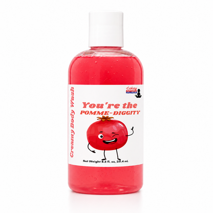 A bottle of Lakay's Remedies 'You're the Pomme-Diggity' creamy body wash, featuring a red liquid inside a clear bottle with a white cap and a white label with a cartoon pomegranate.