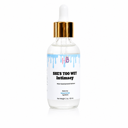 A 2 oz glass dropper bottle of 'She's Too Wet Intimacy' water-based personal lubricant, featuring a white label with blue dripping paint details and a gold collar, set against a plain white background.